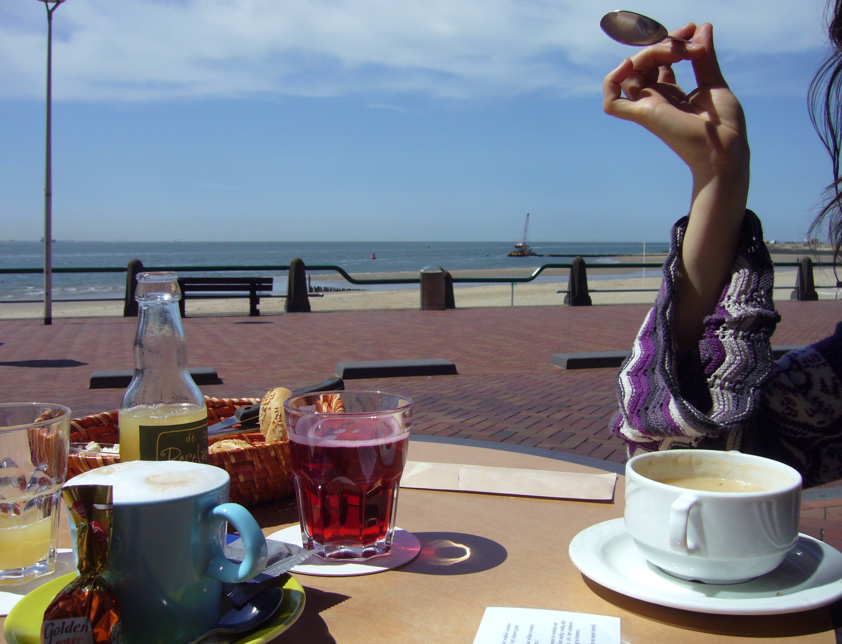 Outdoor café table with coffees, juices, and pastries overlooking a sunny beach promenade and sea