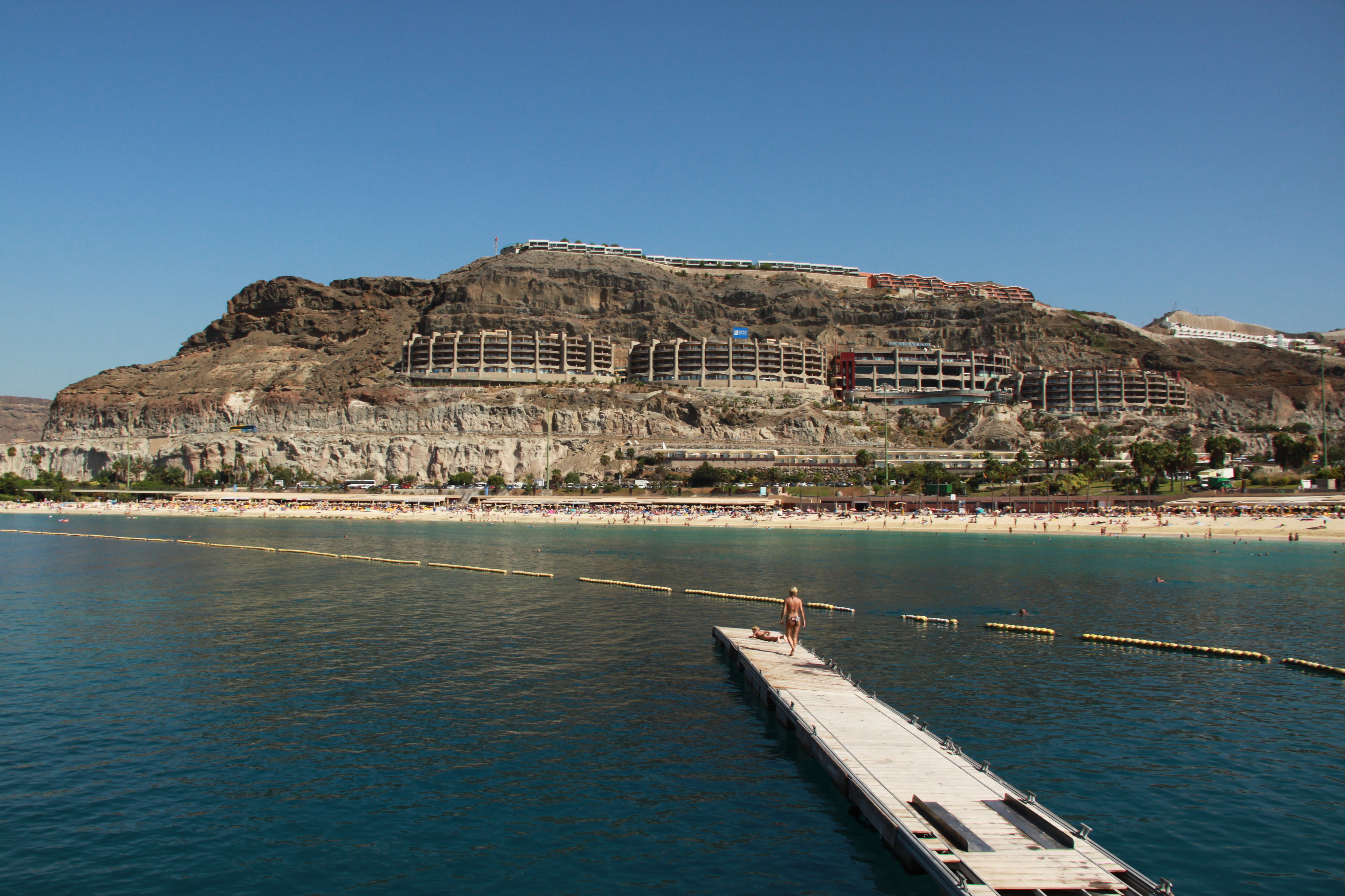 Coastal view of a cliffside resort under construction, beach with umbrellas, and a pier extending into the sea with a person standing on it